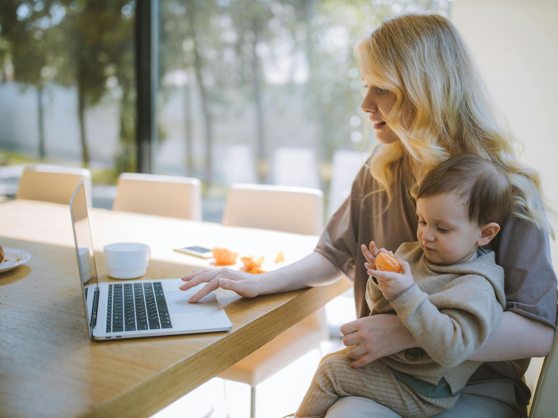 A mother works on a laptop at home while holding her baby, showcasing remote working and parenting.