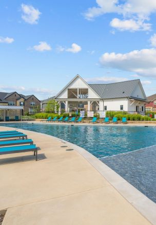Elegant modern house with glass exterior and pool surrounded by lush greenery, under a clear sky.